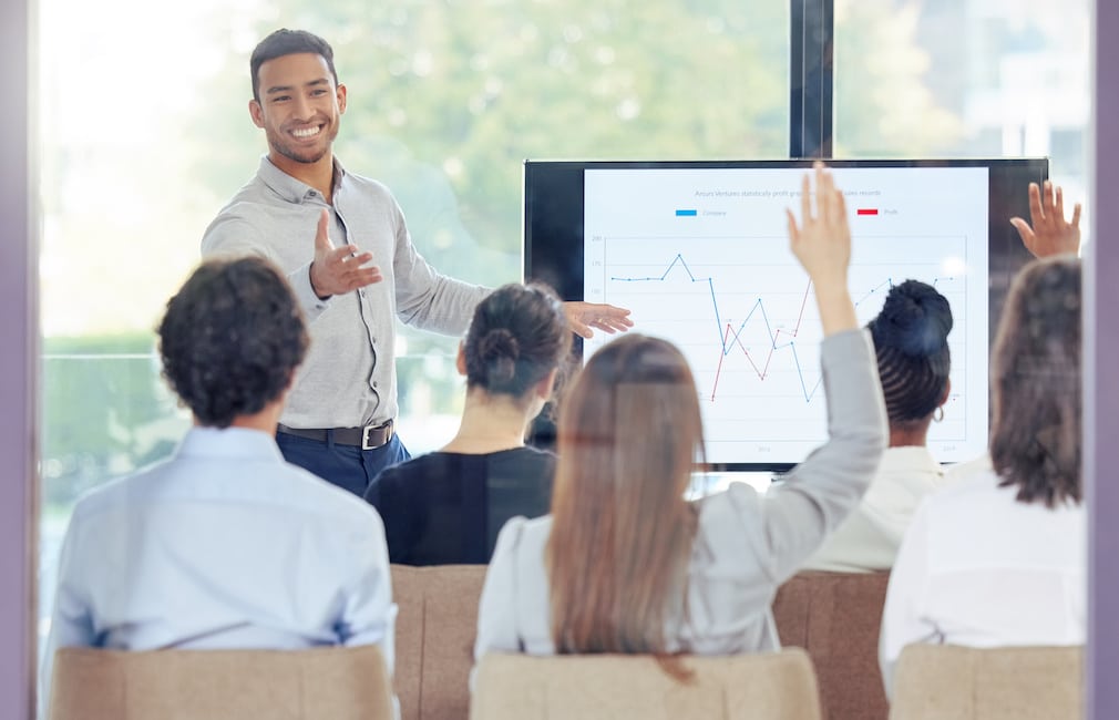 Man at front of a room presenting. A few people have their hands raised.