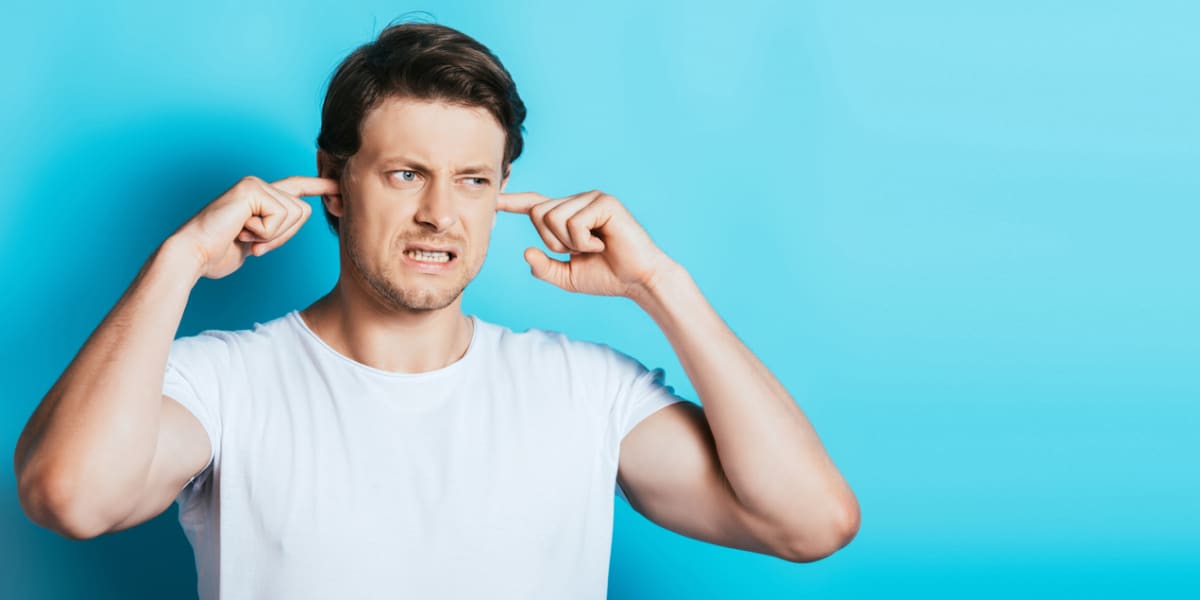 A man in a white t-shirt sticking his fingers in his ears and grimacing.